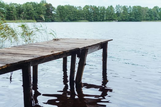 Image of old footbridge made of wooden boards standing on shore of clean lake, offering charming view of green land full of trees and calm water. Tranquil place for relaxation and nature admiration.