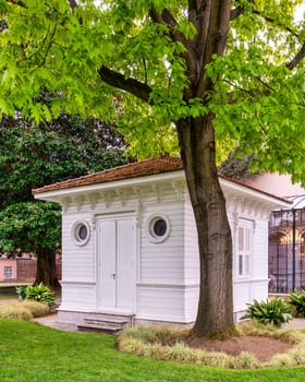White wooden cottage with closed door and shuttered window sits in a rural area. The cottage has a simple rectangular shape and a pitched red bricks roof