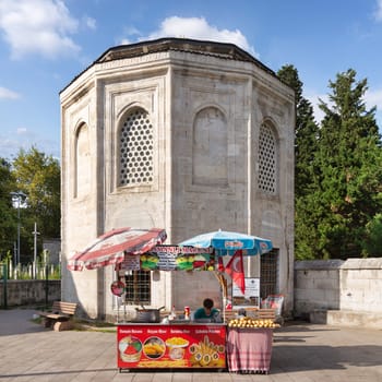 Istanbul, Turkey - September 1, 2022: Young man selling corn, potatoes and ice cream on traditional Turkish fast food cart with historical tomb in the background at Eyupsultan district