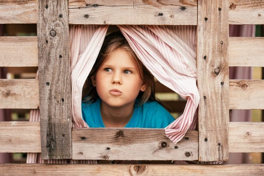 Cute little girl looks out window of wooden children's house. Holidays in village. Happy childhood. Children's games and entertainment. Children's emotions. Positive. Imagination and fantasy.