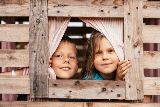 Two charming children, boy and girl, look out small window of wooden children's house. Holidays in village. Happy childhood. Children's games and entertainment. Children's emotions.