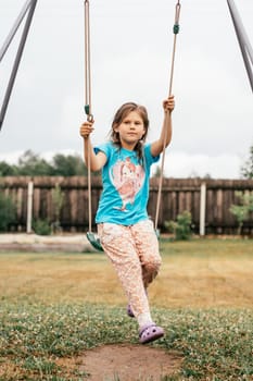 Summer holidays in village. Pretty girl in summer clothes carelessly swings on swing onsunny summer day in nature. Games and entertainment. Summer holidays. Happy childhood. Positive emotions.