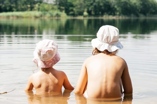 Two children, brother and sister, friends in summer panama hats are sitting in lake in water with their backs to camera. Holidays in village. Happy childhood. Children's games and entertainment.