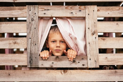 Cute little boy looks out window of wooden children's house. Holidays in village. Happy childhood. Children's games and entertainment. Children's emotions. Positive. Imagination and fantasy.