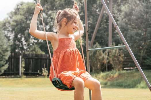 Happy charming little girl in orange sundress is carelessly swinging on swing on sunny summer day in nature. Games and entertainment. Summer holidays. Happy childhood. Positive emotions.