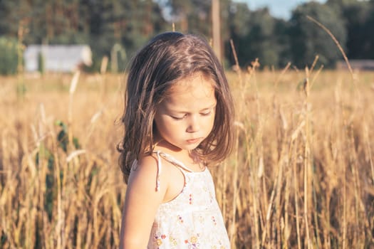 Children in village in summer. Charming little girl in light sundress made of natural fabric on summer day in field among tall cereals. Children's emotions. Happy childhood. Ecological materials.