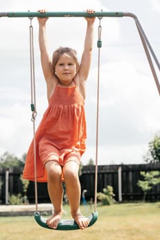 Portrait of lovely little girl in orange sundress standing on swing on sunny summer day. Games and entertainment. Summer holidays. Happy childhood. Positive emotions. World of childhood.