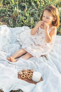 Little beautiful girl in summer colorful sundress has breakfast with gingerbread and milk, sits on white blanket in nature on summer day in village.Natural eco-friendly products. Happy childhood.
