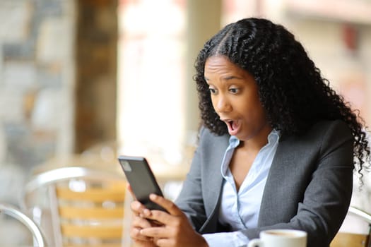 Amazed black executive checking smart phone in a bar terrace