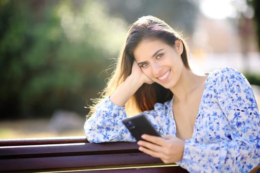Happy woman looks at you holding phone sitting in a park