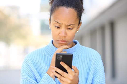 Front view portrait of a worried black woman checking phone in the street