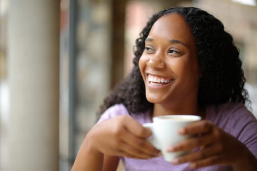 Happy black woman drinking coffee and laughing looking at side in a restaurant terrace
