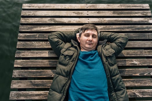 Top view of a man lying on a wooden boardwalk boat dock on a lake with his eyes closed in a jumper and jacket.