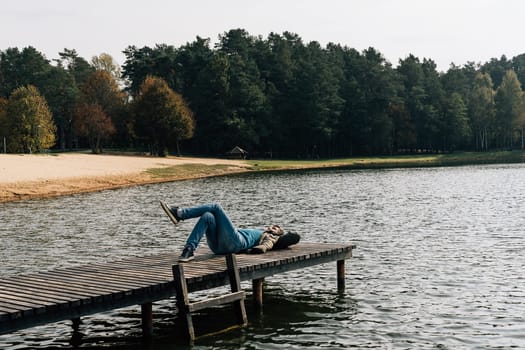 A man rests lying on a wooden pier on a lake in jeans and a jacket on an autumn day.