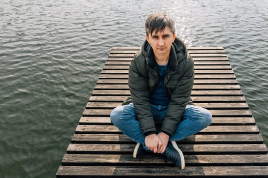 An adult man sits cross-legged on a wooden pier on a lake in jeans and a jacket.