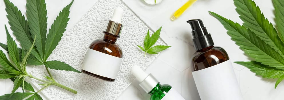 Cosmetic bottles with blank labels near green cannabis sativa leaves on a marble table top view. Mockup, Copy space. Eco friendly CBD skincare beauty products.