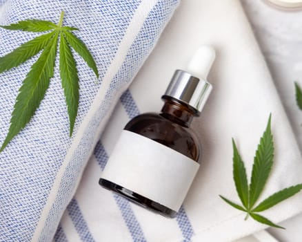 Dropper bottle with blank label near green cannabis leaves on towel top view, mockup. Organic skincare beauty products, alternative CBD cosmetics