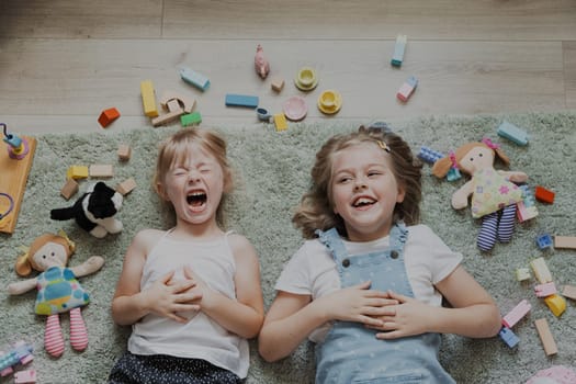 Top view from above on children playing with colorful toys. Portrait of kids lying on carpet at home bedroom or kindergarten. Two happy smiling sisters. Educational eco-friendly games