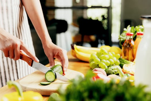 Close up on young housewife woman or girl hands slicing cucumber with kitchen knife, cuts food on wooden chopping board. Unrecognizable female standing at kitchen counter, cutting organic vegetables