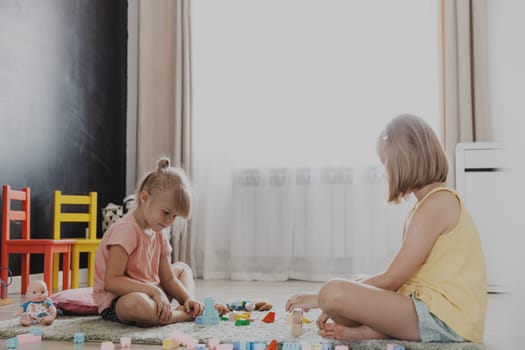 Children playing with colorful wooden toy building blocks, bricks. Kids sitting on floor at sunny white home bedroom or kindergarten, daycare playroom. Funny educational eco-friendly game for child