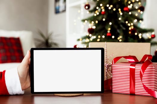 Christmas, New Year background with copy space for design. Santa hand holding empty blank white tablet screen. Mockup for product placement montage. Online shopping from home. Fir tree on background
