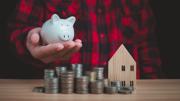 Human holds a piggy bank and there are coins and model wooden houses lined up on the wooden floor. Concepts of finance, savings, investment, and setting goals for fixed deposits with banks.