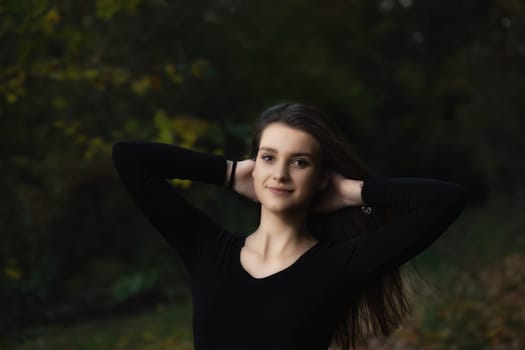 Portrait of attractive long haired young woman posing with hands behind head outdoors on dark background.  Horizontally.