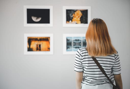 Asian young woman standing she looking art gallery in front of colorful framed paintings pictures on white wall, female watch at photo frame to leaning against at exhibit museum, Back view