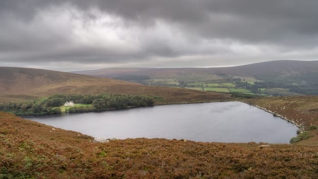 Mansion or house hiding in small forest at the edge of lake Lough Bray, surrounded by dramatic storm clouds. Hiking in Wicklow Mountains, Ireland
