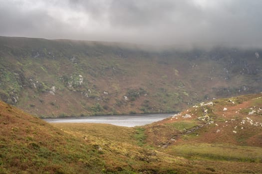 Lake, Lough Bray hiding behind hills and cliffs, surrounded by dramatic storm clouds and sky. Hiking in Wicklow Mountains, Ireland