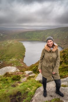 Woman standing on the rock on the top of the mountain and smiling, lake, Lough Bray, down in background. Hiking in Wicklow Mountains, Ireland