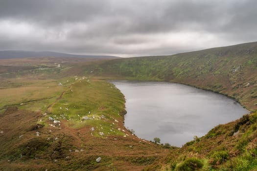 Lake, Lough Bray with mountain trail on the left and cliffs on right, covered in moody, dramatic storm clouds. Hiking in Wicklow Mountains, Ireland