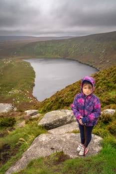 Young girl standing on the rock on the top of the mountain with lake, Lough Bray, down in background. Family hiking in Wicklow Mountains, Ireland