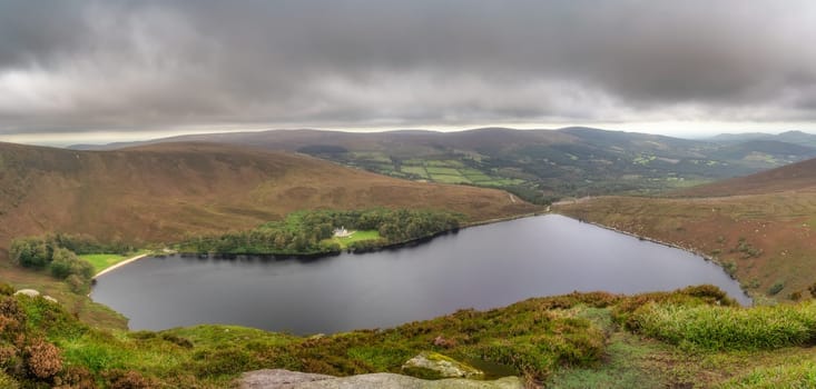 Panoramic view on lake Lough Bray with house, small forest and beach, surrounded by dramatic storm clouds. Hiking in Wicklow Mountains, Ireland