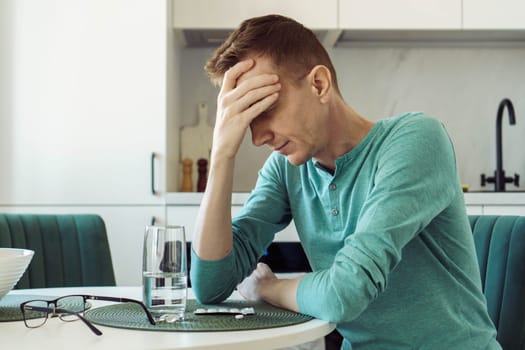 Man in turquoise T-shirt is sitting at home at kitchen table and holding his head with his hand. Illness and loneliness. On table is glass of water and pills, painkillers, antidepressants.