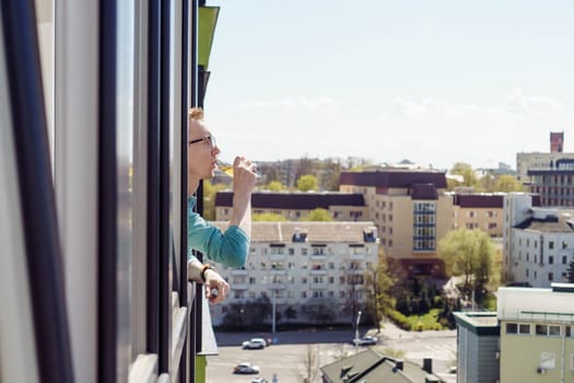 Rest and relaxation. Man with glasses, office worker, freelancer, traveler drinks from glass of juice, beer, drink looks out of apartment window, balcony . City and buildings. Tourism and travel.