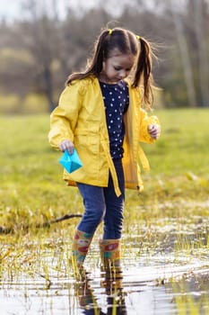 Tranquile child, wearing yellow jacket and rubber boots, standing in puddle with blue paper boat in hand and looking at water, preparing to launch ship. Girl relaxing in nature on fall day. Vertical.