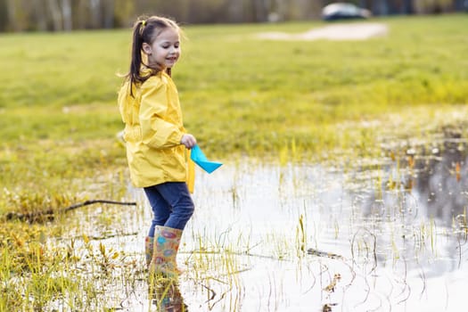 Happy female kid, wearing yellow jacket and rubber boots, standing in puddle on green site, holding blue paper ship in hand and turning head over shoulder with soft smile, enjoying game on fall day.