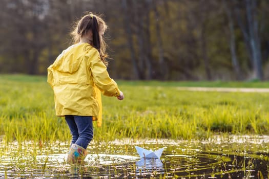 Rear view of little girl, wearing yellow jacket and rubber boots, going in puddle closer to white paper ship, floating on water. Child playing with light ship in autumn day after rain.