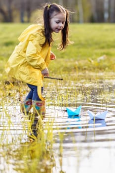 Active little girl in yellow jacket and rubber boots standing in puddle and looking at two paper ships of different colours, floating on water. Kid watching light boats, playing in nature. Vertical.