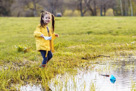 Portrait of gorgeous little girl, adorned in yellow jacket and rubber boots, holding white paper ship in hand and looking at camera with shiny smile, preparing to launch boat on puddle in autumn.