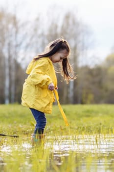 Wonderful little girl, wearing yellow jacket and rubber boots, standing in puddle surrounded by green lawn and looking down at water with curiosity, spending autumn day in nature. Vertical.