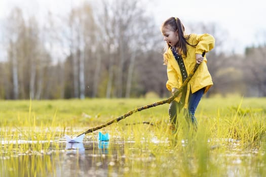 Amazed female kid in yellow jacket and rubber boots, standing in puddle on green lawn and looking at two paper boats with open mouth. Little girl playing with ships using long branch on fall day.