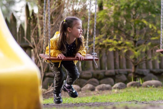 Thoughtful female child, wearing yellow jacket, hanging on swings at playground and looking away, distracted from relaxation. Girl with two ponytails holding board of seesaw and flying through air.