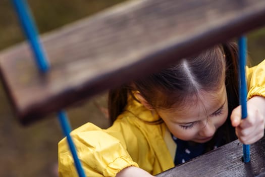 Active little lady, wearing bright yellow jacket, carefully climbing on wooden staircase built at playground, holding blue rope and looking ahead, relaxing and having fun on fresh air on autumn day.