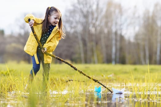 Charming little girl, adorned in bright yellow jacket and rubber boots, holding long branch in hands while standing in puddle and moving two papers ships of various colors, floating on water.