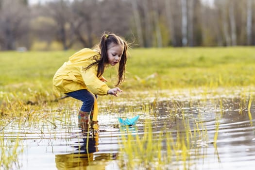Charming little girl in yellow jacket and rubber boots standing in puddle on green lawn, stretching arm forward and bending over, carefully looking at blue paper boat, floating on water.
