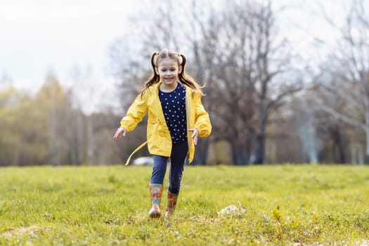 Loveable little girl with two ponytails, wearing yellow jacket and rubber boots, strolling across green site on autumn day, looking down with smile and eyes full of curiosity and carefree.