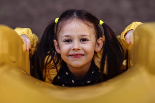 Portrait of charming little girl with brown hair tied up in two ponytails looking at camera and smiling while lying on stomach on yellow slide, having delightful walk at playground on autumn day.