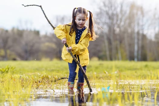 Concentrated little girl, wearing yellow jacket and rubber boots, holding long branch in hands while standing in puddle and looking down with focused eyes, preparing for playing with paper boats.